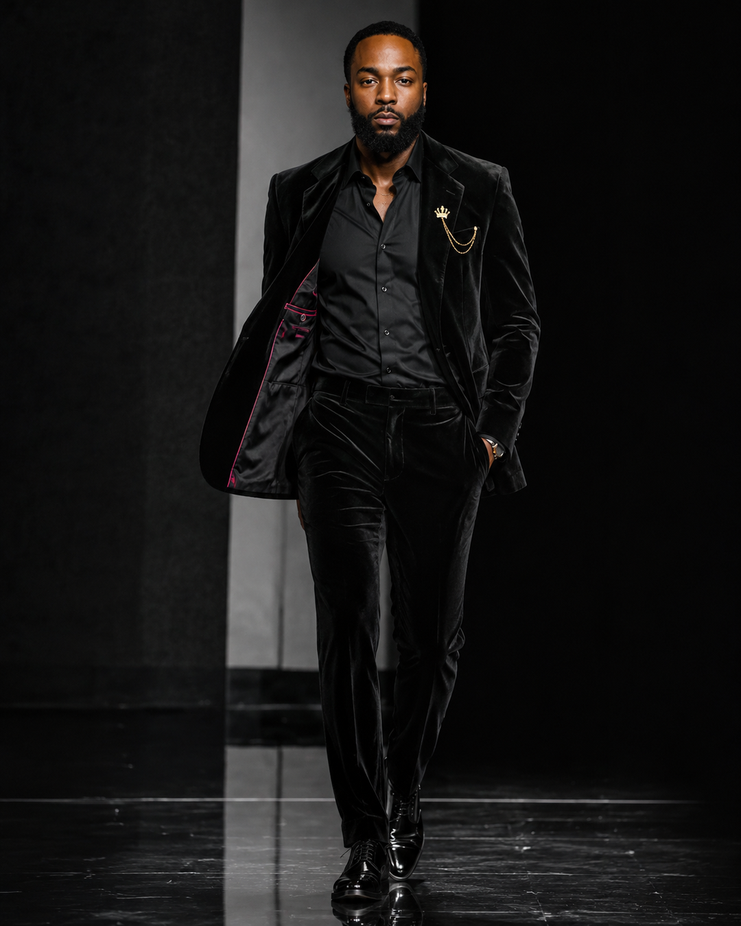 A male model walks down a runway wearing a black velvet suit with a black shirt, gold chain lapel pin, and pink lining visible on the jacket interior.