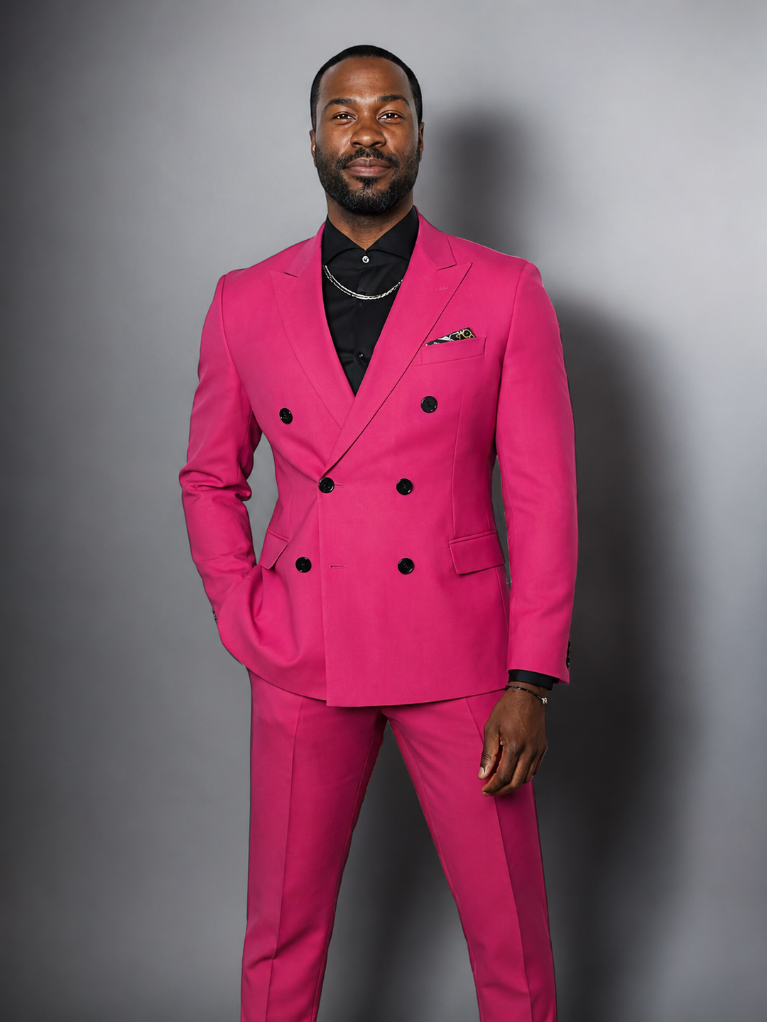 A man wearing a bright pink double-breasted suit with black buttons, a black shirt, and a silver chain necklace, standing against a dark gray studio background.