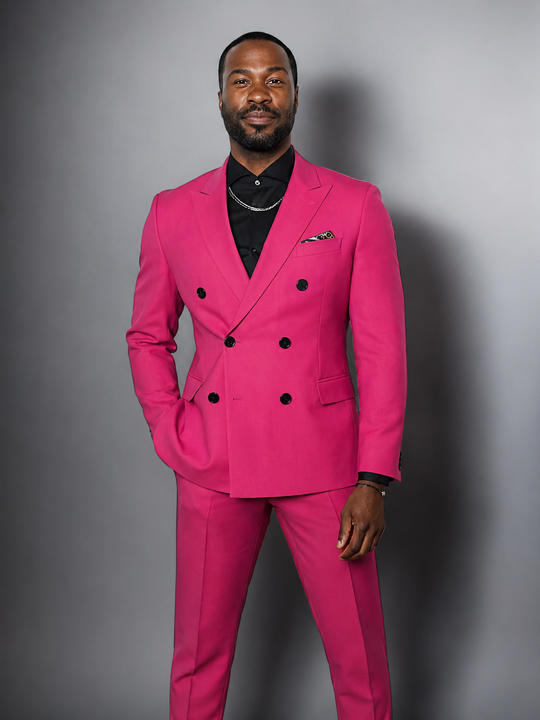 A man wearing a bright pink double-breasted suit with black buttons, a black shirt, and a silver chain necklace, standing against a dark gray studio background.